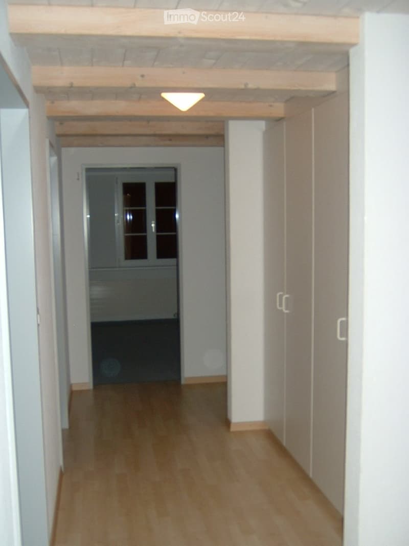 Empty hallway with wooden flooring, white walls, and a ceiling light fixture. A door with glass panels is visible at the end of the corridor.