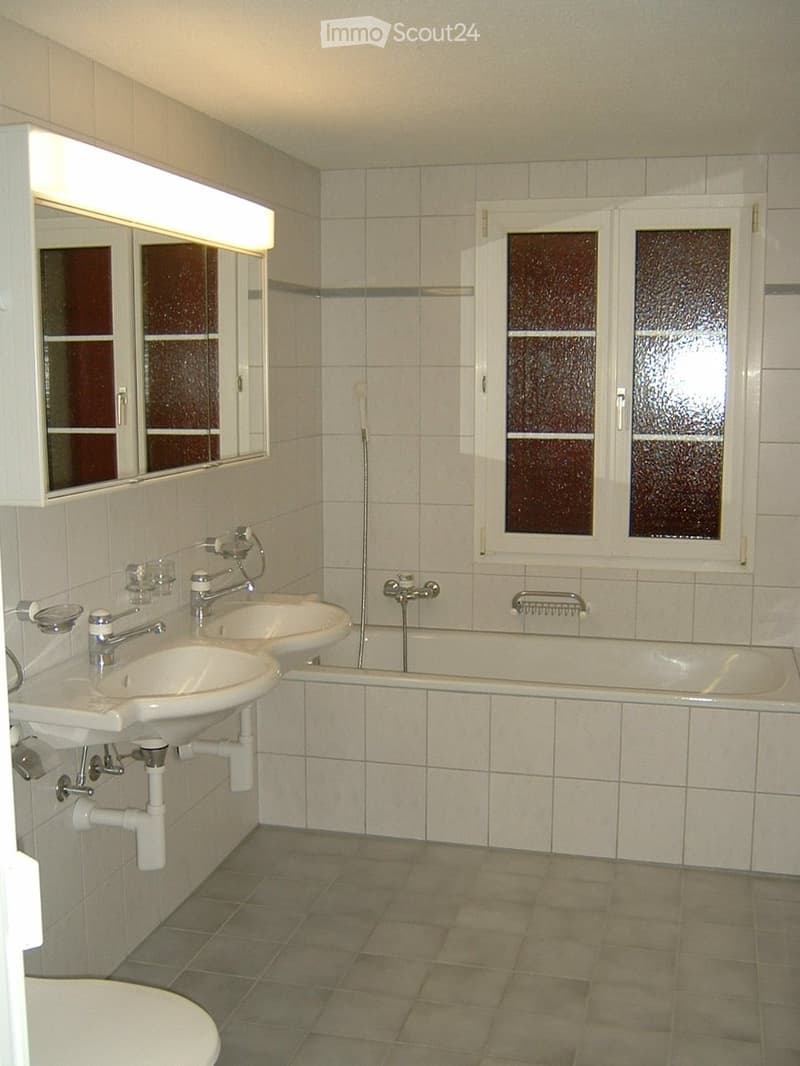 Modern bathroom with two sinks, a bathtub, a toilet, and a large mirror with built-in lighting. The room features tiled walls and floor, and frosted glass windows.