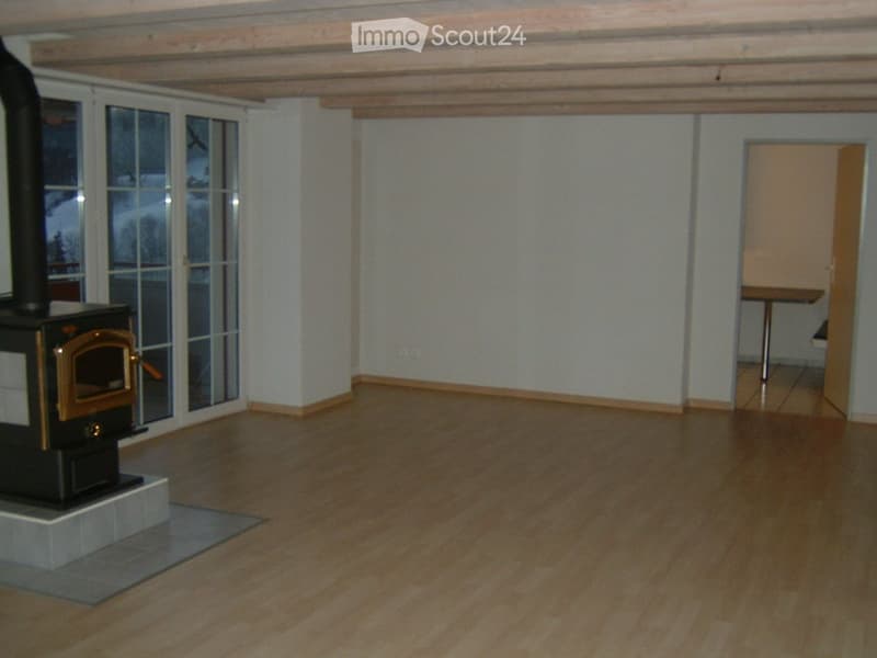 Empty room with wooden flooring, white walls, and a wood-burning stove. Large windows with a view of the outside, and a doorway leading to another room.