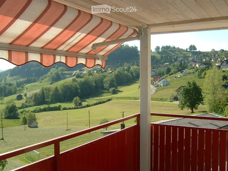 Balcony with red wooden railing, striped awning, overlooking a scenic countryside with green fields, scattered houses, and distant hills.