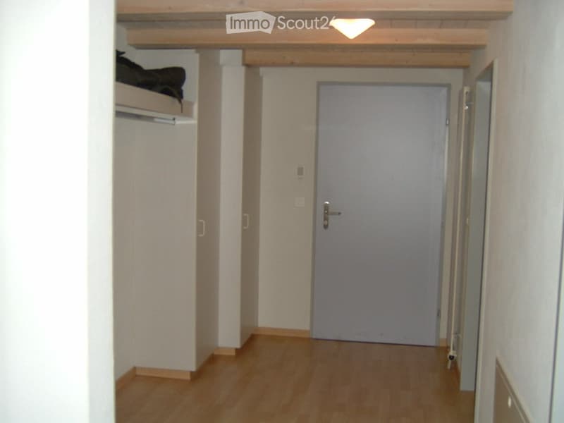 A narrow hallway with wooden flooring, white walls, and a closed door at the end. A built-in closet with a bunk bed is visible on the left side.