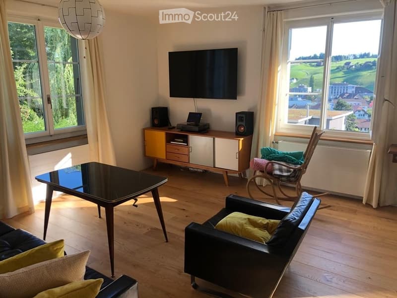 Modern living room with large windows offering a scenic view, featuring a black leather sofa, a glass coffee table, a wooden TV stand, and a rocking chair.