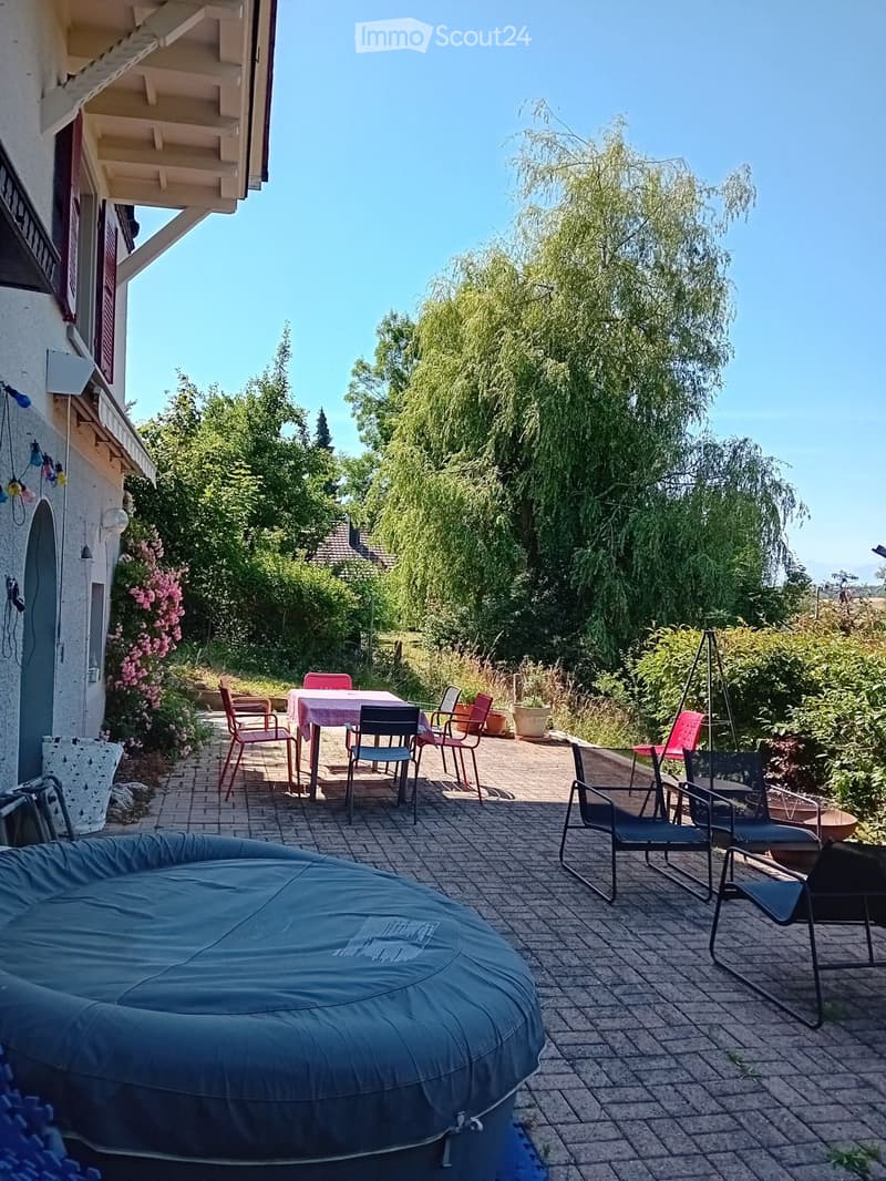 Outdoor patio with brick flooring, a round sofa, a table with chairs, and surrounding greenery.