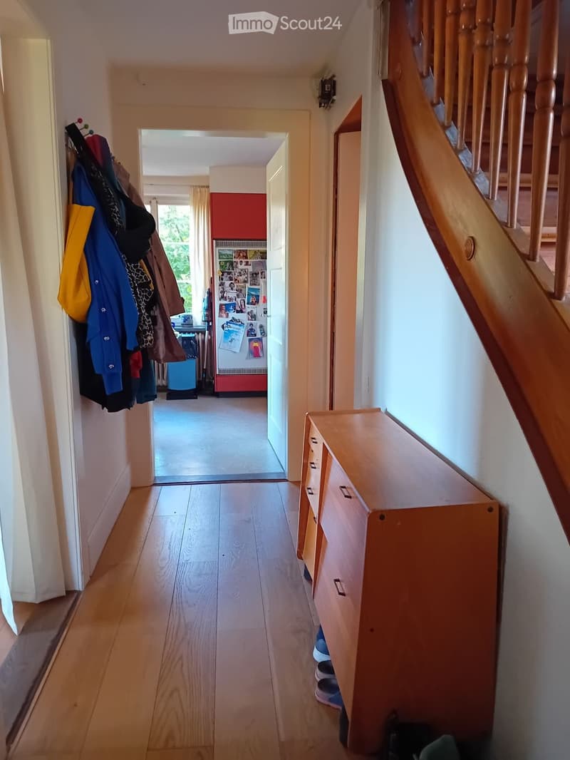 A well-lit hallway with wooden flooring, a staircase on the right, and a coat rack on the left. An open doorway leads to another room with a red accent wall and visible furniture.