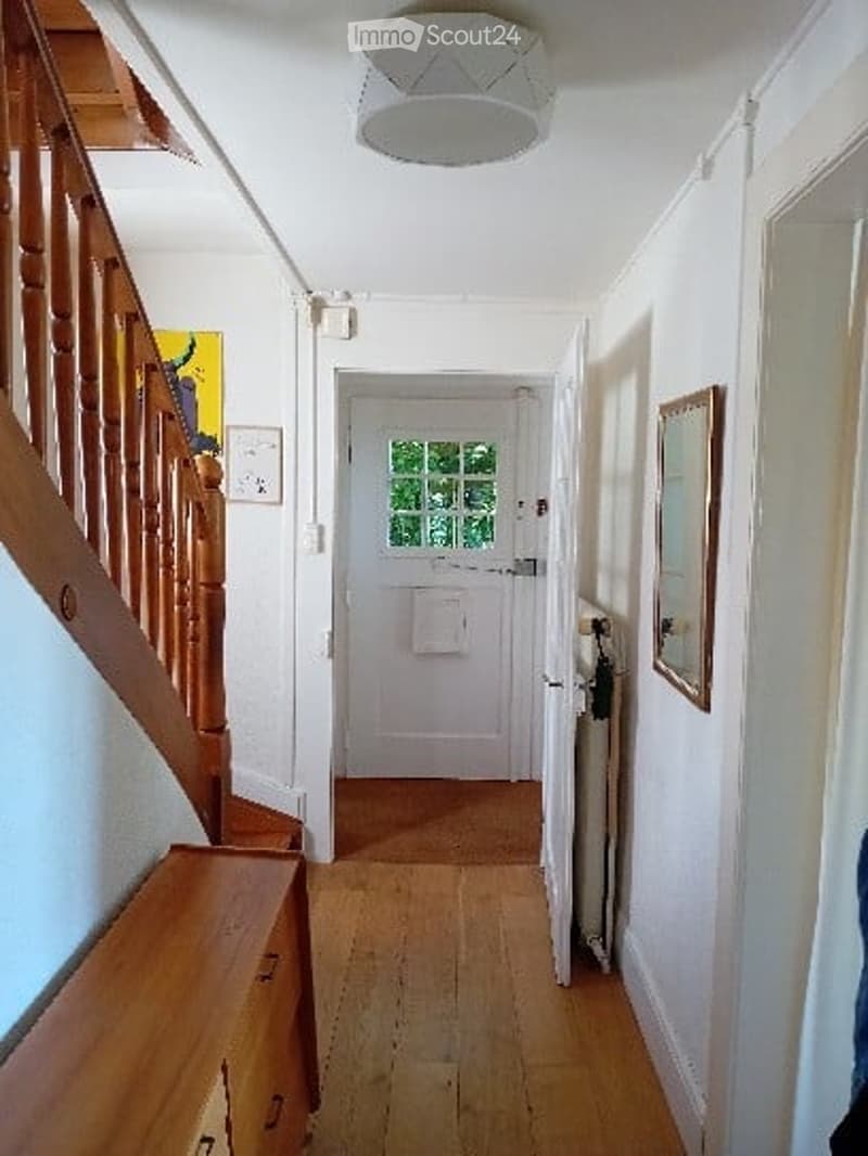 A well-lit hallway with wooden flooring, a staircase on the left, and a front door at the end. The walls are painted white, and there is a framed picture on the right wall.