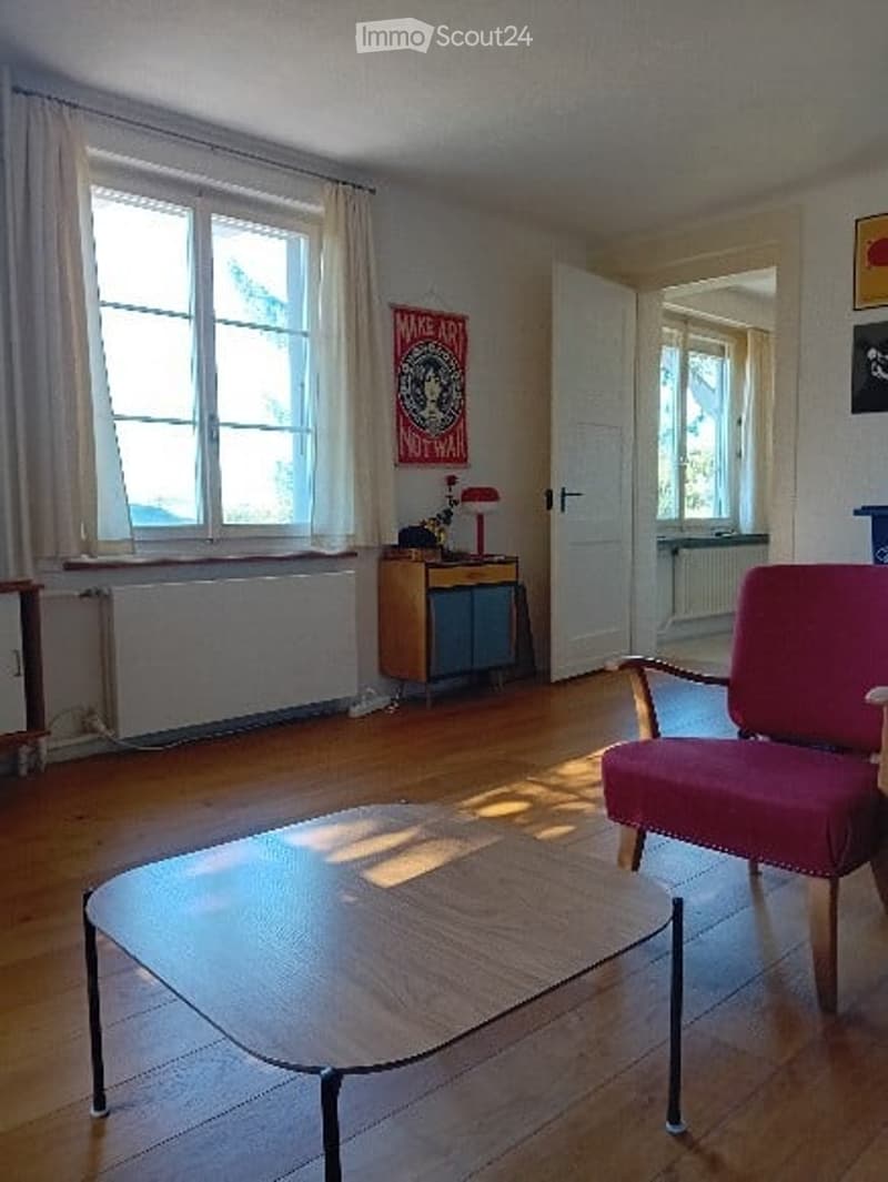 Bright living room with wooden flooring, large windows, a red armchair, a coffee table, a side table with a lamp, and a radiator under the windows.