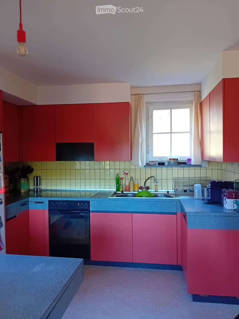 Modern kitchen with red and pink cabinets, a central island, and a window with curtains. The kitchen features a sink, oven, and various kitchen items on the counter.