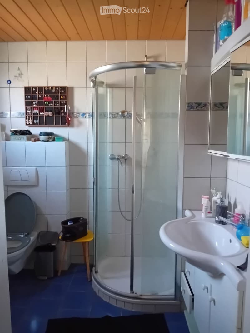 Bathroom with a toilet, sink, shower, and various toiletries on the sink counter. A small stool holds a bag, and a cabinet is visible.