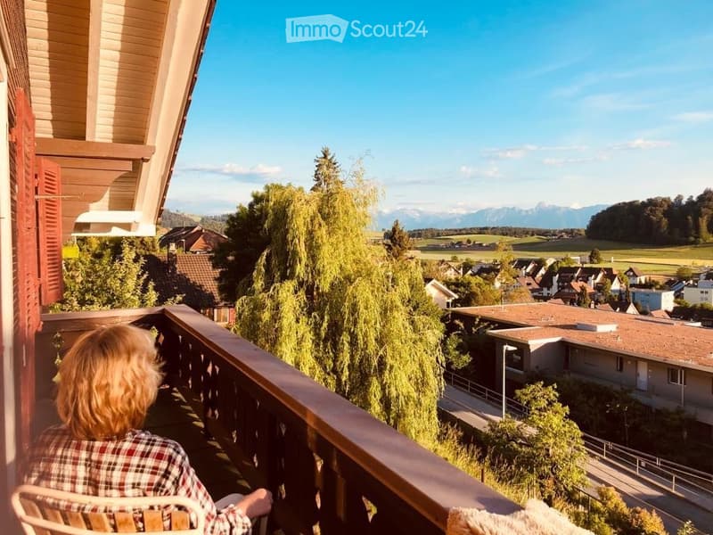 Balcony overlooking a scenic valley with houses, trees, and distant mountains under a clear blue sky.