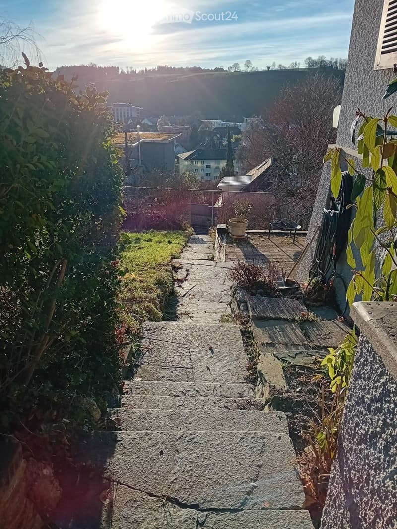 A stone staircase leads up to a small outdoor area with a wooden deck, potted plant, and a view of a town with houses and greenery in the background.