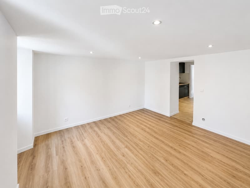 Empty room with light wooden flooring and white walls, featuring recessed ceiling lights and an open doorway leading to a small kitchen area.