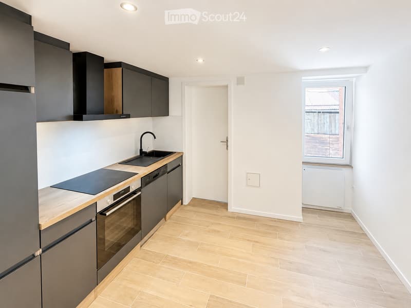 Modern kitchen with light wooden flooring, sleek black and grey cabinets, stainless steel appliances including a refrigerator, oven, dishwasher, and sink, and a window offering a view of the outdoor area.