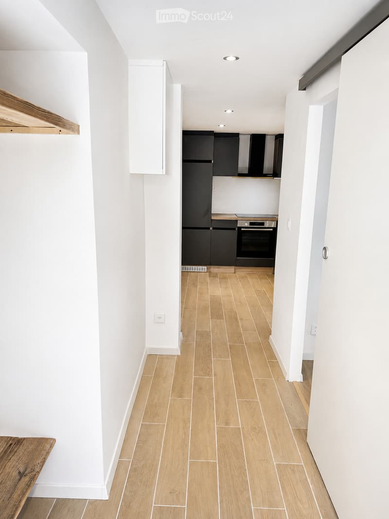 Bright, modern hallway with light wooden flooring, white walls, and recessed ceiling lighting. Features a small wooden bench on the left and a doorway leading to a kitchen area with dark cabinets and stainless steel appliances.