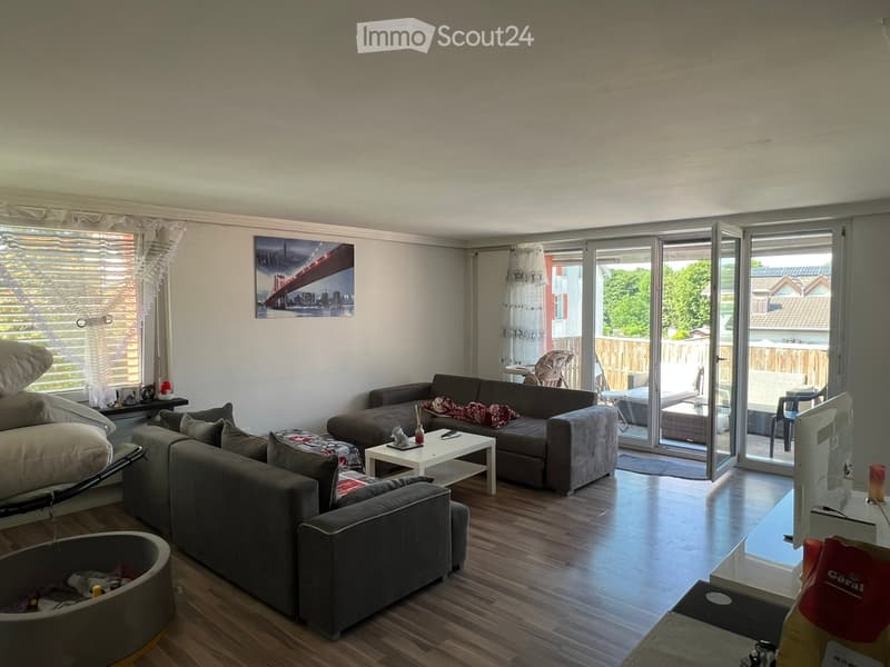 Modern living room with wooden flooring, large sliding glass doors leading to a patio, a grey sectional sofa, a white coffee table, and a TV screen on a stand.