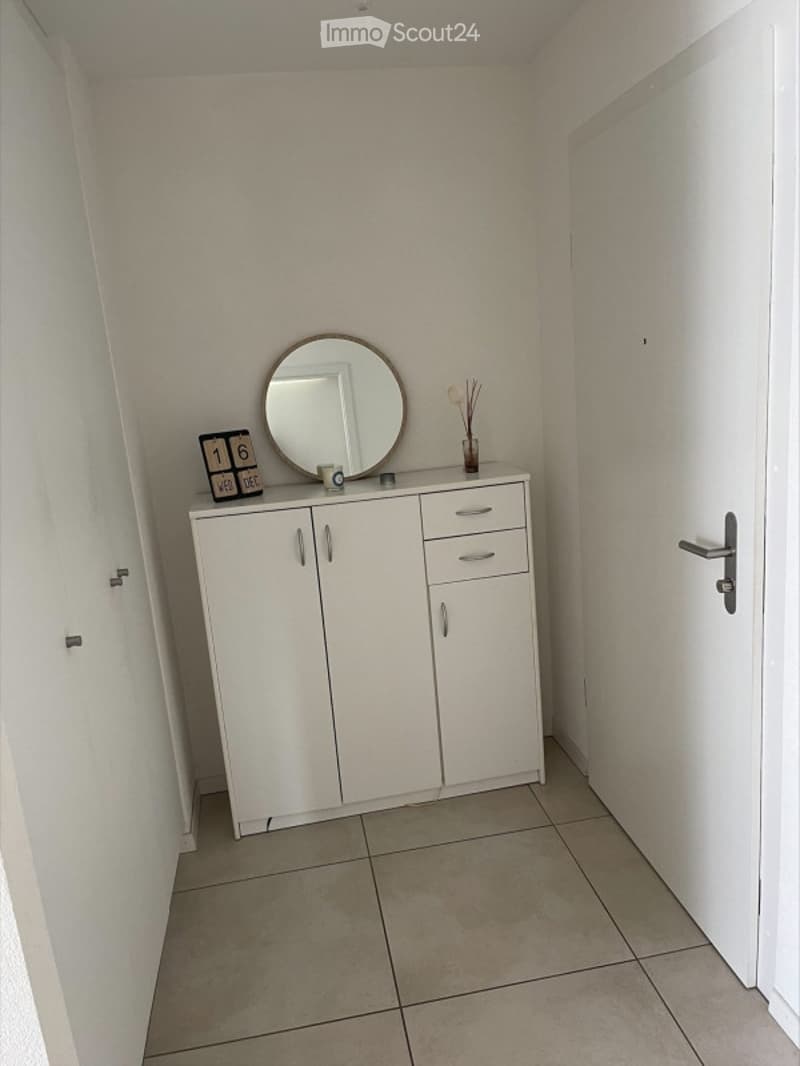 A small, neatly arranged hallway or entryway with a white cabinet, a round mirror, a decorative vase, and a small clock on top of the cabinet.