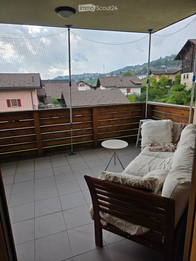 Balcony with wooden railing, tiled floor, sofa with cushions, small round table, and a view of surrounding houses and hills.