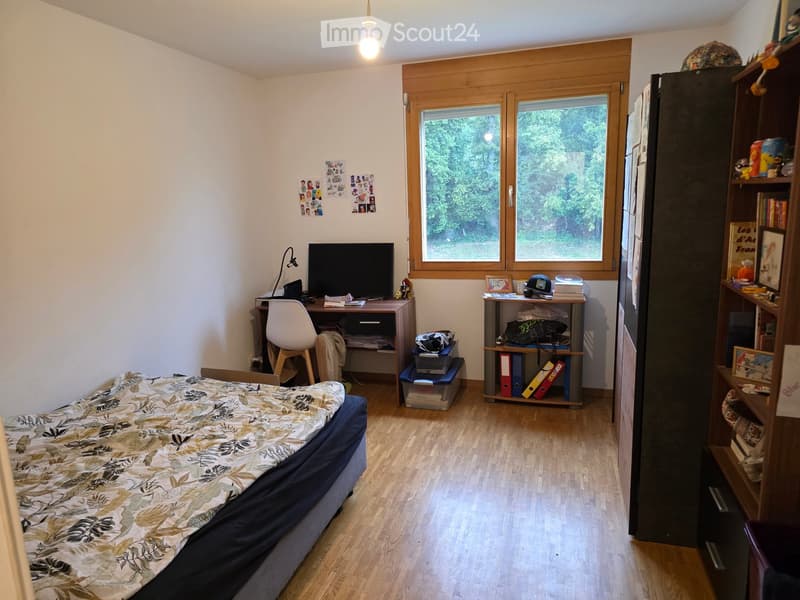 A well-lit bedroom with a bed, desk, chair, and shelves. The room features wooden flooring, large windows with a view of greenery, and various personal items including books and decorations.