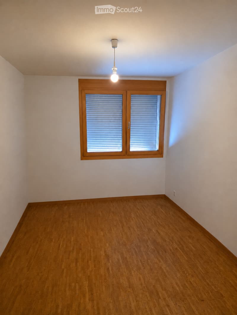 Empty room with wooden flooring, white walls, and a single window with blinds. A pendant light fixture hangs from the ceiling.