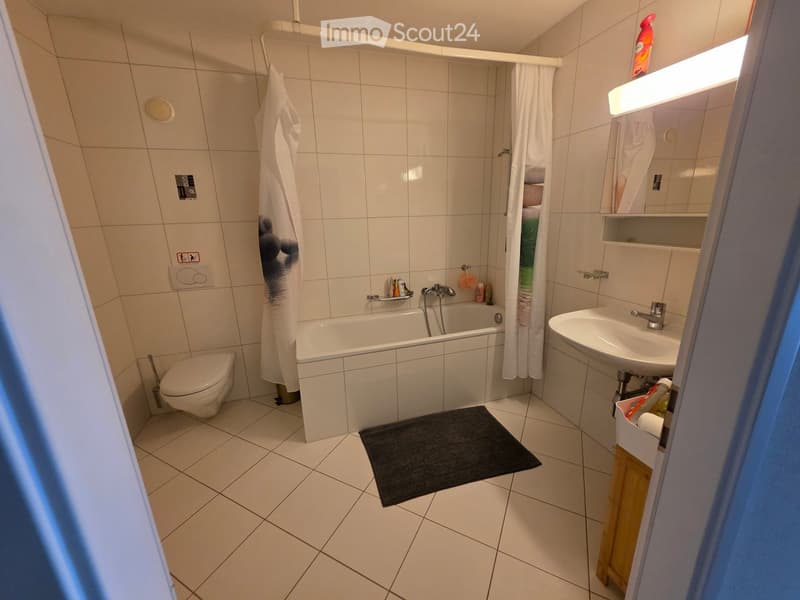 Modern bathroom with white tiles, bathtub, toilet, sink, mirror, and a black bathroom rug on the floor.