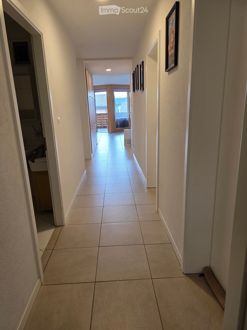 A clean, tiled hallway with white walls, recessed lighting, and several framed pictures on the right wall. The hallway leads to an open area with a view of a balcony or outdoor space.
