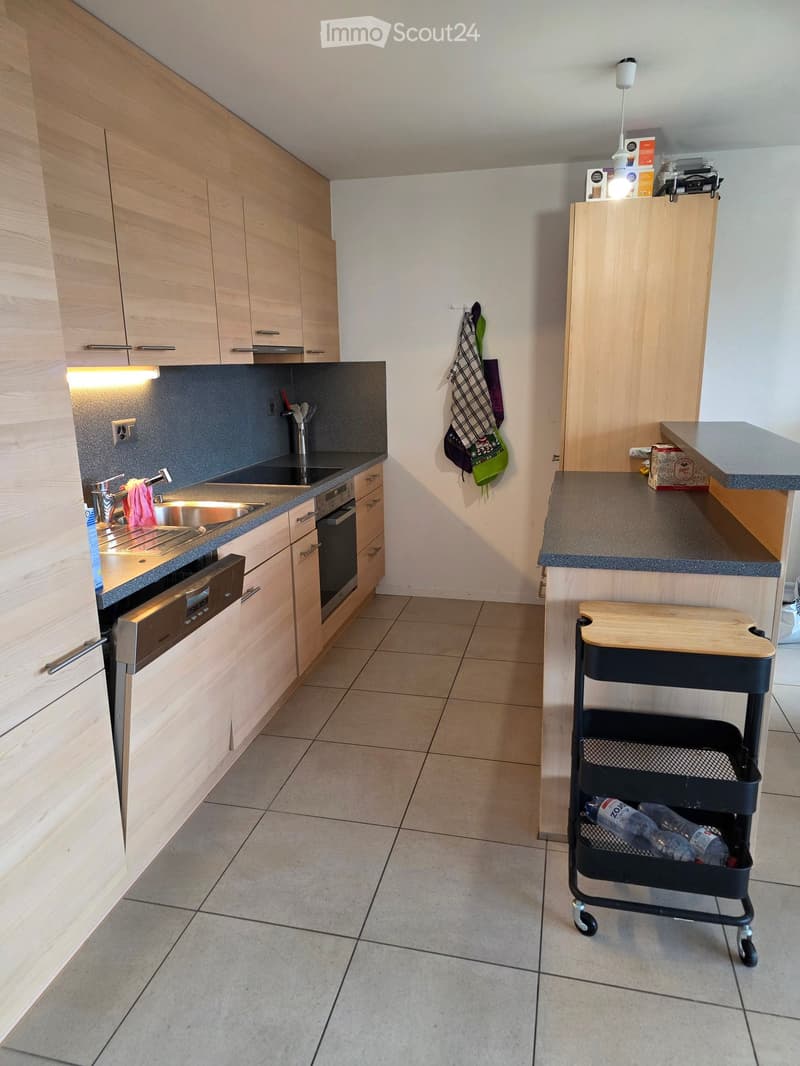 Modern kitchen with light wooden cabinets, stainless steel sink, oven, and a small cart with a wooden cutting board on top.