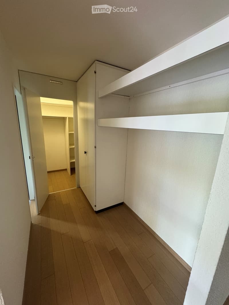 Empty hallway with wooden flooring, white walls, built-in shelving, and an open closet door leading to another room.