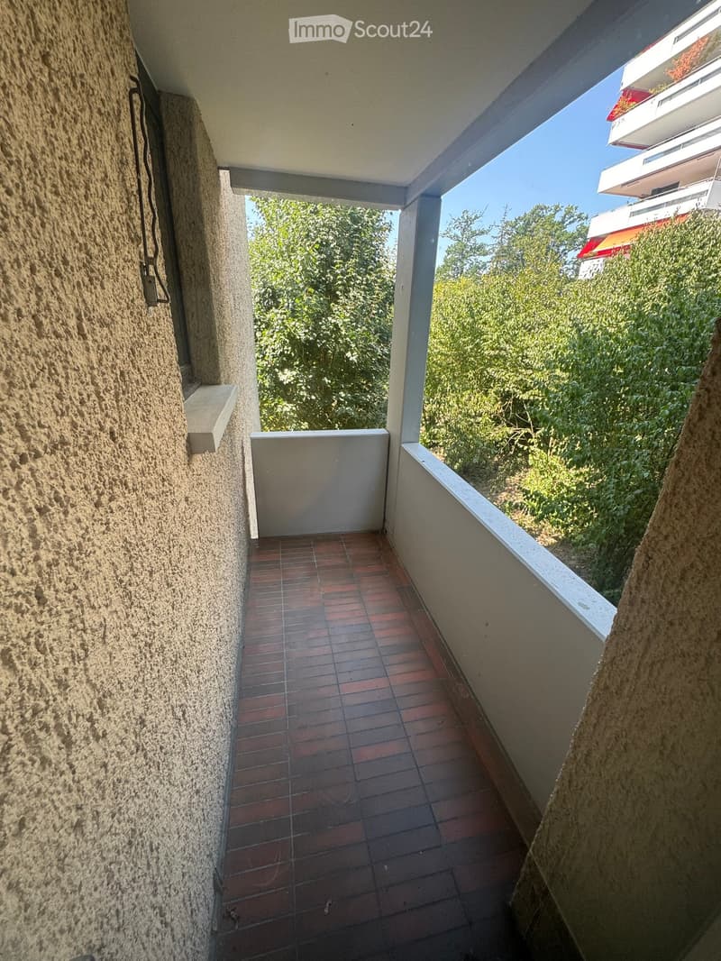 Balcony with red tiled floor, white railings, and a view of greenery and neighboring buildings.