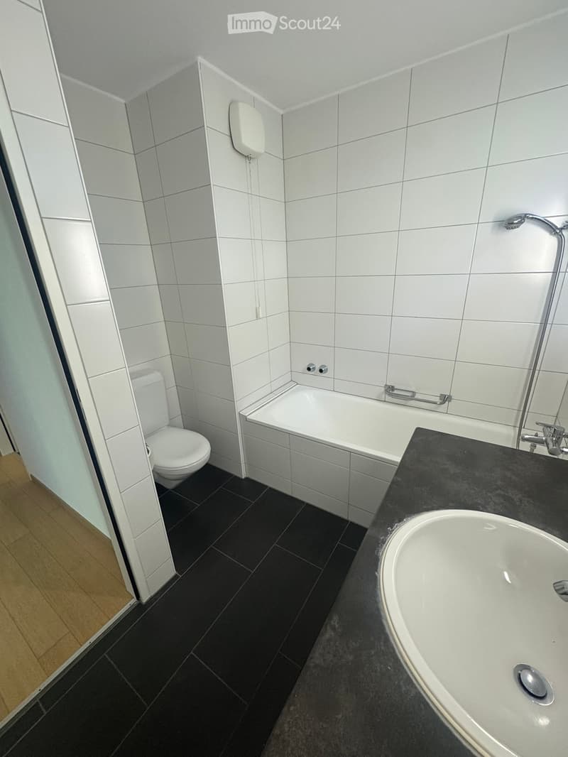 Modern bathroom with white tiles, black floor tiles, a toilet, a bathtub, and a sink with a faucet.