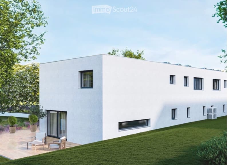 Modern white house with large windows and a raised ground floor, surrounded by a well-maintained lawn and small garden area with potted plants and outdoor seating.