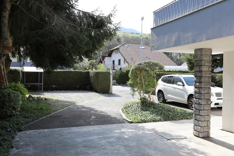 Driveway with a car parked, concrete floor, hedges, trees, building with a balcony