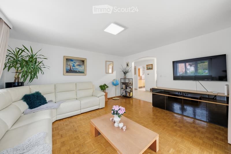 Modern living room with white walls, wooden flooring, a large sectional sofa, a coffee table with a flower arrangement, a TV mounted on the wall, and a bookshelf with decorative items.