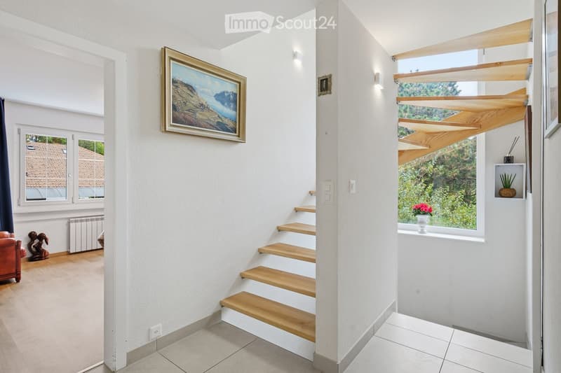Bright hallway with wooden stairs, white walls, large windows, and a painting on the wall.