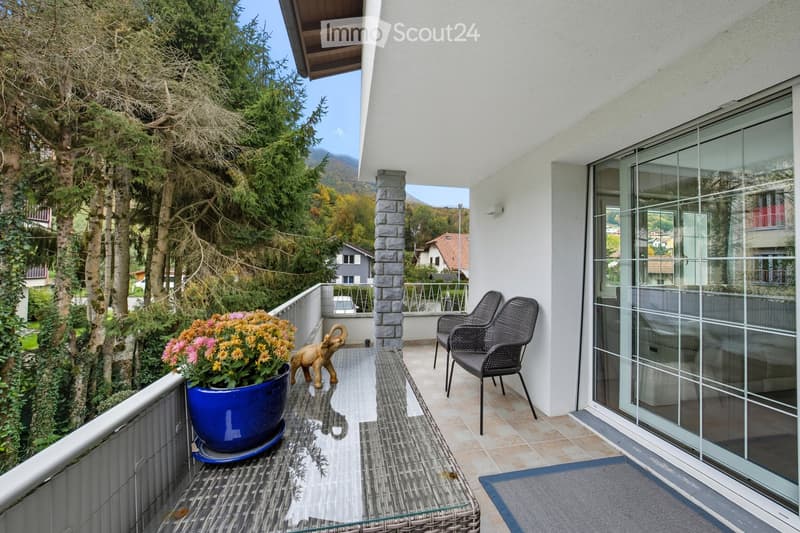 Balcony with a view of trees and mountains, featuring a wicker chair, a glass table, a large blue flower pot with flowers, and a small elephant statue.