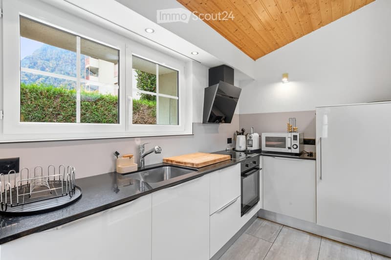 Modern kitchen with white cabinets, stainless steel appliances, and a wooden cutting board on the counter. The kitchen features a sink, microwave, and a view of greenery outside.