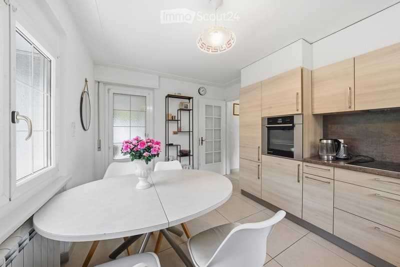 Modern kitchen with light wooden cabinets, stainless steel appliances, a round white dining table with chairs, a vase of pink flowers, and a small shelf with decorative items.