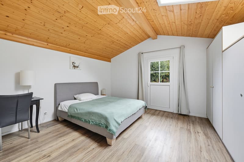Cozy bedroom with a wooden slanted ceiling, a grey upholstered bed with a green throw, a small black desk with a chair, and a white lamp on a side table.