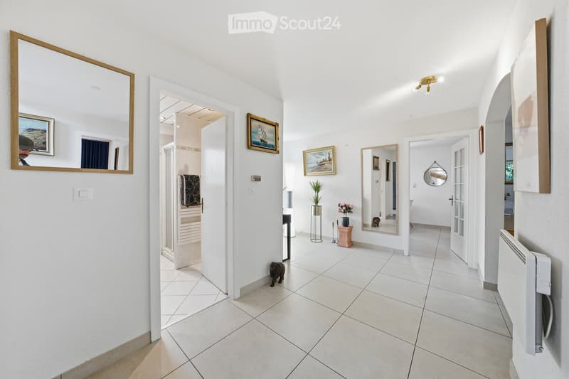 Bright hallway with white tiled flooring, white walls, and several doors leading to different rooms. Features include a large mirror, framed artwork, small decorative items, and a small statue on the floor.