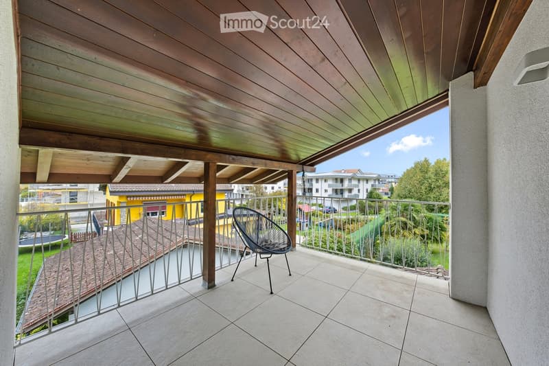 Balcony with wooden ceiling, tiled floor, metal railing, and a single modern chair. Offers a view of the surrounding area with buildings and greenery.