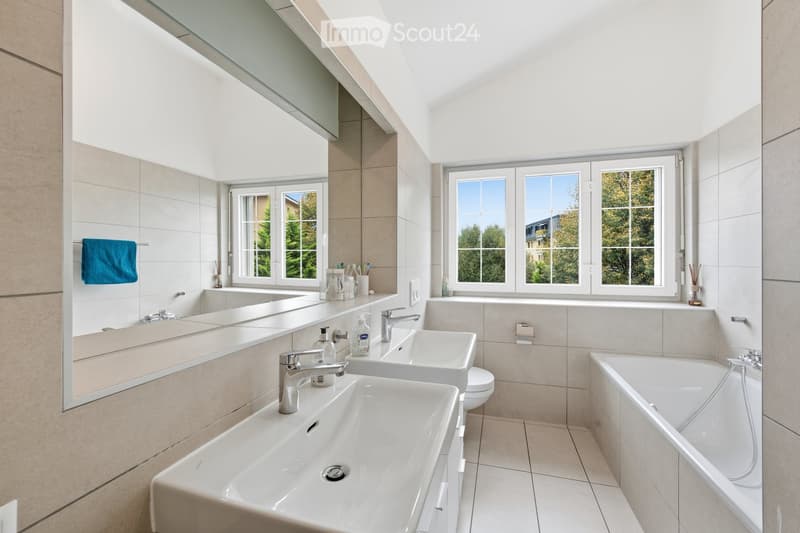 Modern bathroom with two sinks, a bathtub, a large mirror, and a towel hanging on the wall. The room features large windows with a view of greenery outside.