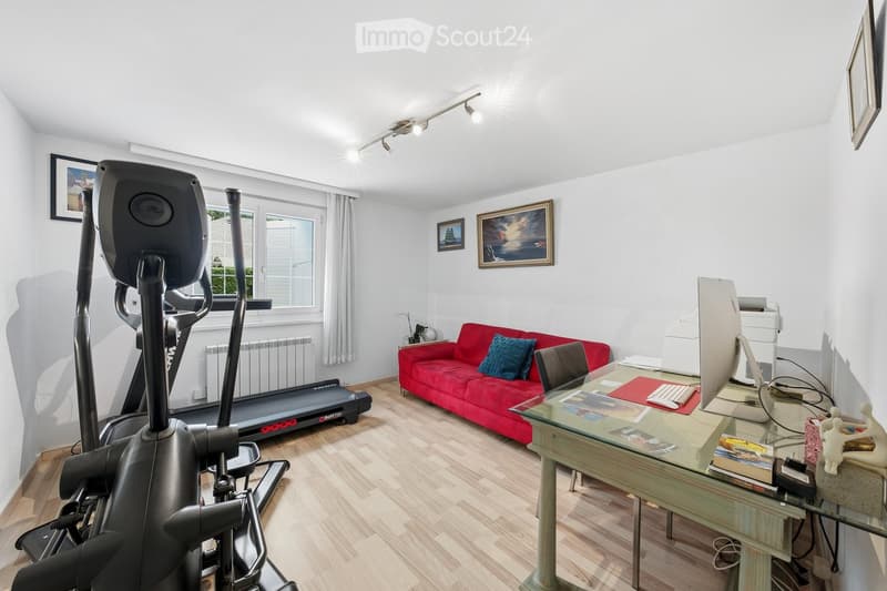 A well-lit room with wooden flooring, a red sofa, a treadmill, an exercise bike, a glass table with a keyboard and screen, and framed pictures on the walls.