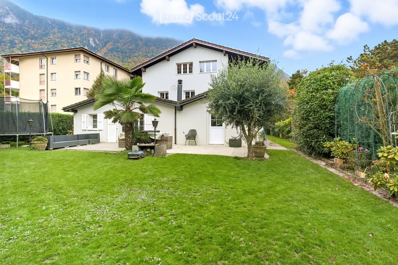 Well-maintained garden with a lawn, patio seating area, outdoor furniture, and surrounding greenery, including trees and shrubs. A white house with multiple windows and a chimney is visible in the background, along with a neighboring building and a trampo