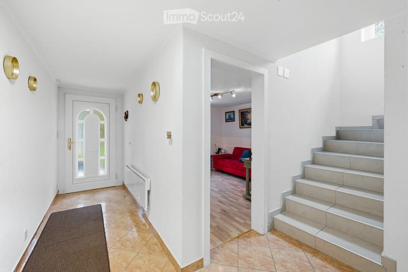 Bright hallway with white walls, tiled floor, and a staircase leading upwards. Features a red sofa visible in the adjacent room.