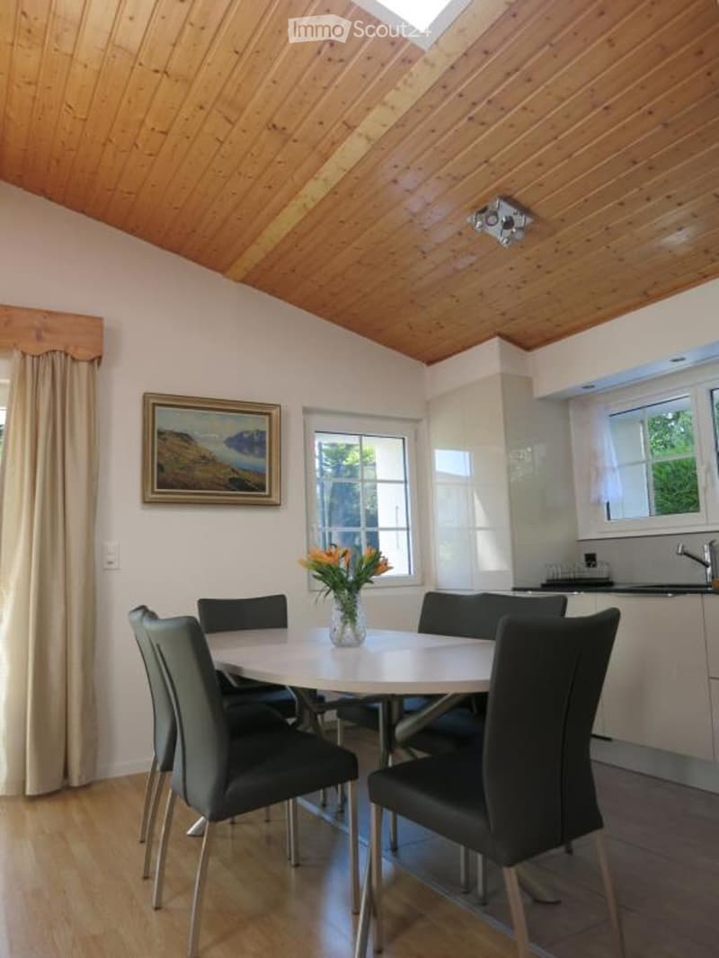 Bright dining area with a round white table surrounded by black chairs, a vase with orange flowers, and a modern kitchen area with white cabinets and a sink.