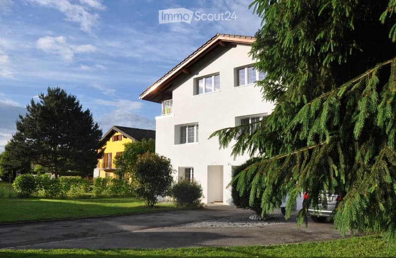 2 story house, white exterior, brown roof, balcony, driveway, car parked, trees, bushes, yellow house in background