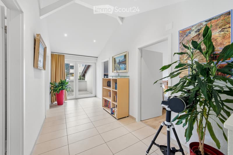 Bright hallway with white walls, tiled floor, large potted plants, bookshelf, and framed artwork. Features a sliding glass door leading to a balcony area.