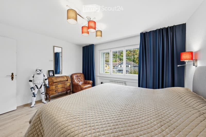 Well-lit bedroom with a large bed, leather armchair, wooden dresser, mirror, and wall-mounted lamp. The room features dark blue curtains and a view of the outdoors through large windows.
