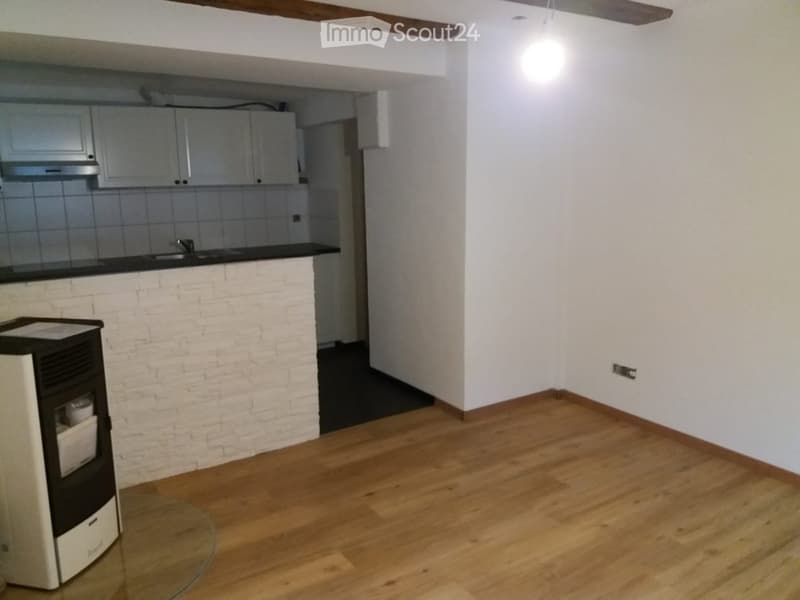 A modern, unfurnished living space with wooden flooring, white brick accent wall, and a small kitchen area featuring cabinets, a sink, and a refrigerator.