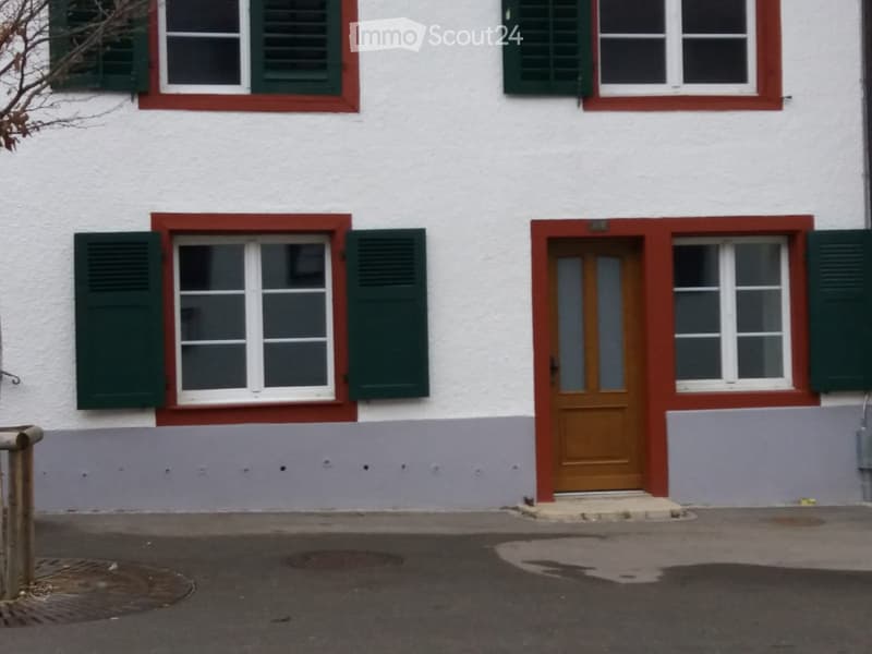 Two-story building with white walls, red trim around windows and door, green shutters, and a brown front door.