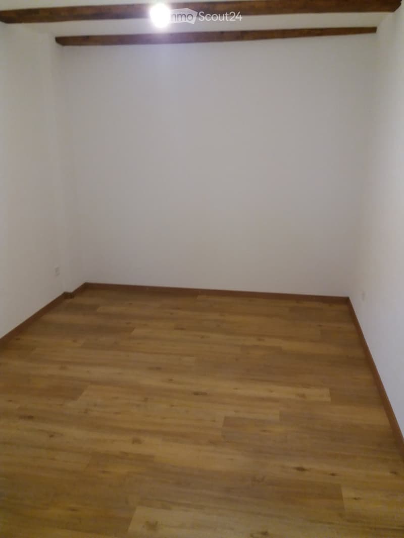 Empty room with wooden flooring and white walls, featuring two wooden beams on the wall and a light fixture.