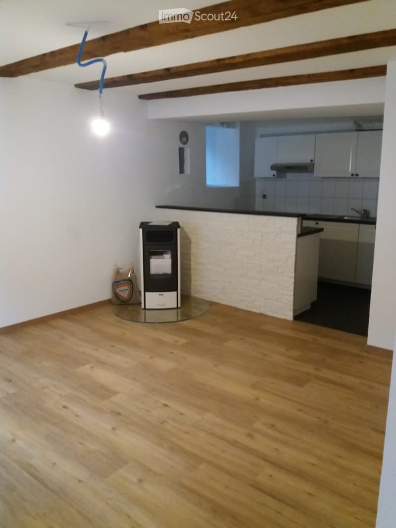Empty room with wooden flooring, white brick accent wall, exposed wooden beams on the ceiling, and a small kitchen area visible in the background.